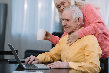 senior woman holding coffee cup and hugging man sitting at table and using laptop