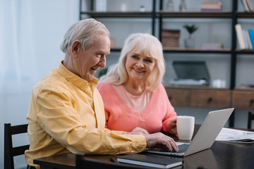 senior couple in colorful clothes sitting at table with coffee cup and using laptop
