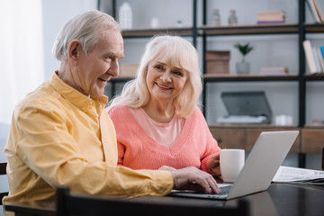 smiling senior couple in colorful clothes sitting at table and using laptop