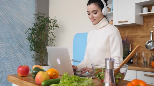 Smiling Young Woman Using Laptop In Kitchen Wearing Headphones At Home