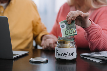 Fototapeta premium cropped view of senior couple putting money in glass jar with 'pension' word