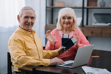 senior man looking at camera and holding air tickets with passports near surprised woman at home