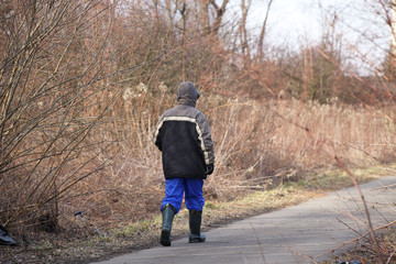 A worker in rubber boots and a jacket walks along an asphalt road past the brushwood in the springtime. Formation of green spaces and the landscape design. Unemployed looking for a job