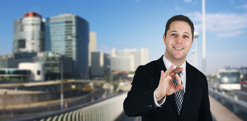 Fototapeta na wymiar Businessman Smiling and Feeling Happy Giving Ok Sign With Business City and Corporate Buildings In Background