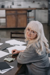 Fototapeta na wymiar sad female pensioner sitting at table, looking at camera and counting money at home