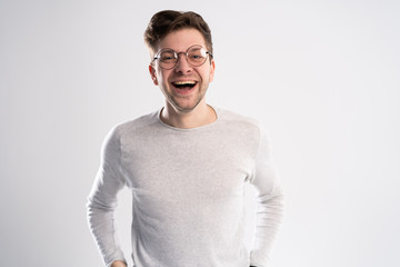 Portrait of young Caucasian guy enjoys funny story or joke, laughs joyfully, has toothy smile, wears white t shirt in one tone with background. Happiness and youth concept.