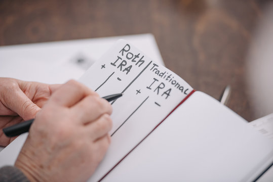 Cropped View Of Senior Woman Writing In Notebook With Roth Ira And Traditional Ira Words