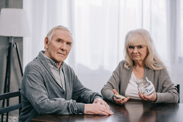 sad senior couple sitting at table, looking at camera and holding empty glass jar with 'pension' lettering