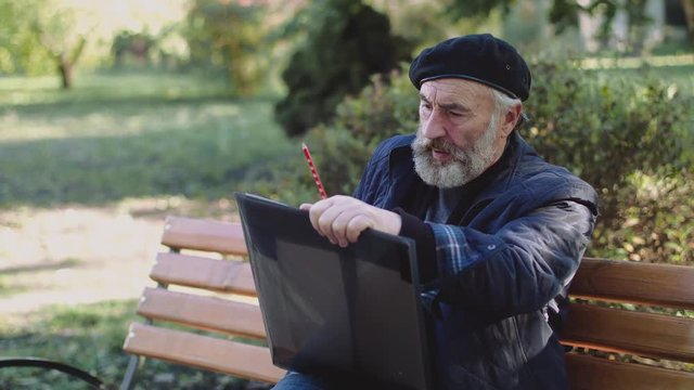 Wrinkled artist sits on street bench, begins smoking and drawing picture