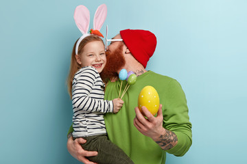 Fototapeta na wymiar Happy father and daughter wears bunny ears, holds painted eggs, celebrate Easter together, kiss and hug, enjoys spring holiday, appreciate family traditions, have last preparations. Feast day