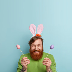 Vertical shot of optimistic redhead male laughs joyfully, has fun during painting Easter eggs, prepares for important spring holiday, enjoys festive event with family, isolated on blue wall.