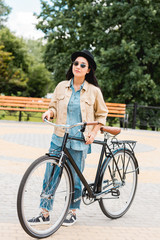 cheerful girl in sunglasses and hat standing with bicycle in park