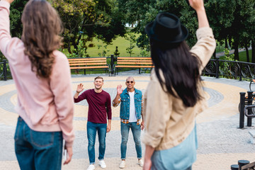 Fototapeta premium selective focus of cheerful young multicultural men waving hand and smiling to female friends in park