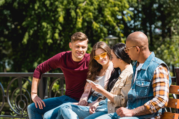 Fototapeta na wymiar cheerful multicultural friends sitting on bench and looking at girl holding smartphone