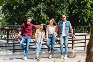 Fototapeta premium Cheerful multicultural young men standing with longboard and American football near girls