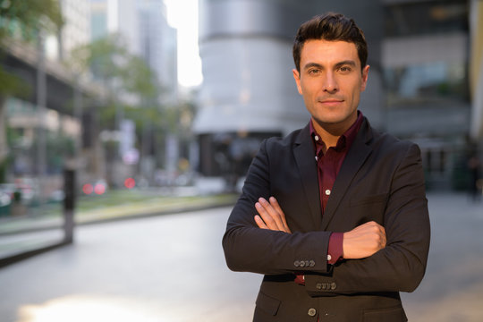 Young handsome Hispanic businessman with arms crossed outside the office building - Powered by Adobe