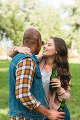 Fototapeta premium cheerful girl whispering in ear of african american boyfriend while holding bottle