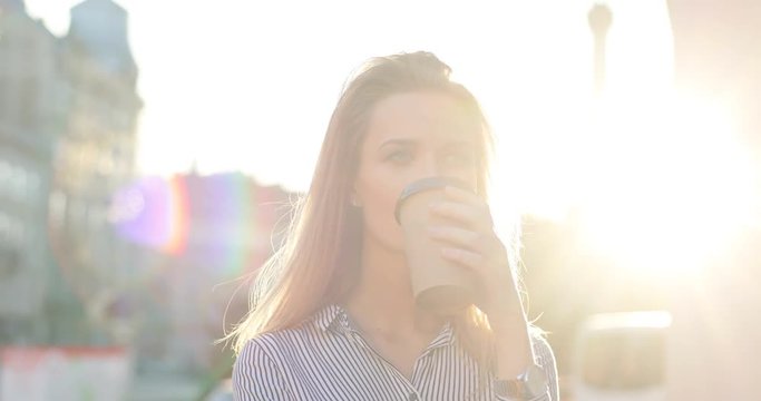 A beutiful girl is drinking her morning coffee-to-go in the city centre and smiling. The sun is shining at the camera. Front view, close-up.