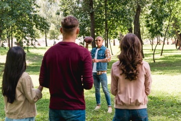 Fototapeta premium back view of friends looking at african american man holding american football in park