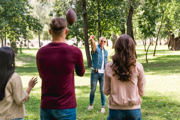 back view of friends looking at african american man throwing american football in park - Powered by Adobe