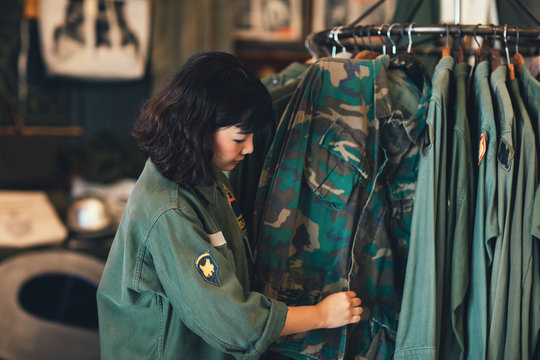 Pretty Asian woman standing by the clothes rack at vintage military shop and folding shirts.