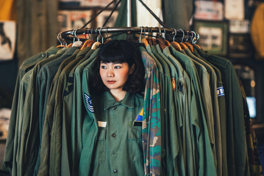 Portrait Of Asian Woman Shop Assistant Sitting By The Clothes Rack At Vintage Military Shop.