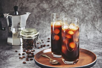 Ice coffee in a tall glass with cream poured over and coffee beans on a old rustic wooden board. Cold summer drink on a dark wooden background with copy space 