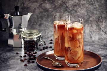 Ice coffee in a tall glass with cream poured over and coffee beans on a old rustic wooden board. Cold summer drink on a dark wooden background with copy space 