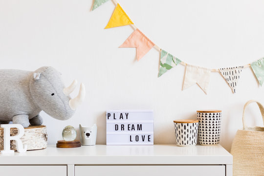 The modern scandinavian newborn baby room with plush rhino, design boxes, mock up blackboard and natural basket. Hanging cotton flags on the white background wall. Stylish and cozy interior.