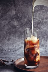 Ice coffee in a tall glass with cream poured over and coffee beans on a old rustic wooden board. Cold summer drink on a dark wooden background with copy space 