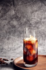Ice coffee in a tall glass with cream poured over and coffee beans on a old rustic wooden board. Cold summer drink on a dark wooden background with copy space 