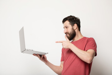 Portrait of a joyful young man in red t-shirt looking at laptop computer and celebrating isolated over grey background 