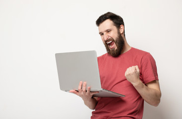 Portrait of a joyful young man in red t-shirt looking at laptop computer and celebrating isolated over grey background 