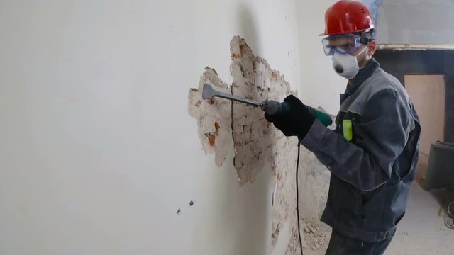 worker in protective suit demolishes plaster wall. Dirty, hard work. Personal protective equipment. Helmet, respirator and goggles.