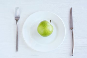 Juicy organic green apple on white plate with selective focus and blurred fork and steel knife on neutral wooden background. Diet concept with healthy vegetarian food for weight loss