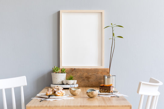 Stylish and modern interior design of kitchen space with small wooden table  with mock up photo frame, avocado plant, succulents, cups of tea and tasty dessert. Scandinavian space decor.