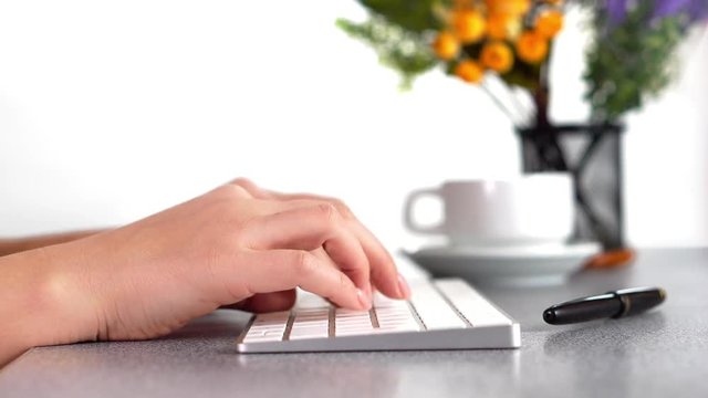 Closeup video of female hands, typing on the PS keyboard, at the table in the office. Flowers in the background.