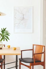 Fototapeta na wymiar Stylish and modern dining room interior with mock up poster map, sharing table design chairs, gold pedant lamp and cups of coffee. White walls, wooden parquet. Tropical leafs in vase. Eclectic decor.