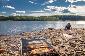 summer  landscape by the river white clouds on the blue sky.  white fish steak is fried on the grill. in the background is a teenage girl in a white t-shirt sitting on the river Bank.