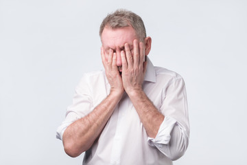 Senior man in white shirt covering his face with both hands