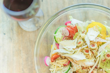 Bowl of fresh cheese vegetable salad and glass of red wine placed on wooden table.