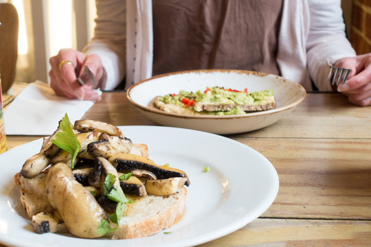 Woman Eating Helathy Avocado On Toast With Dish Of Mushrooms On Toast In Foreground