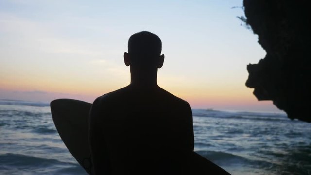 Closeup silhouette of a man of average build at sunset, with a surfboard in his hands, who looks into the distance at sea