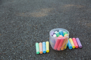 Plastic transparent box full of new colorful chalks outdoors isolated on surface of grey grunge pavement as background. Horizontal color photography.