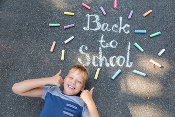 Back to school concept. Inscription on pavement of sidewalk at city street made with colorful crayons by young kid. Portrait of boy with thumbs up. Horizontal color photography.