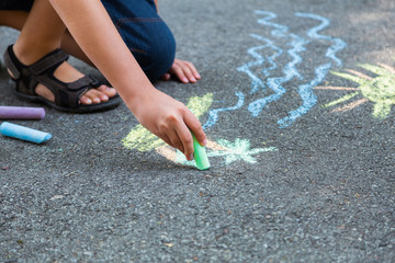 Closeup top view of child's little hand holding piece of blue chalk and drawing sea water on pavement of sidewalk outdoors at city park. Child drawing tropical beach. Horizontal color image.