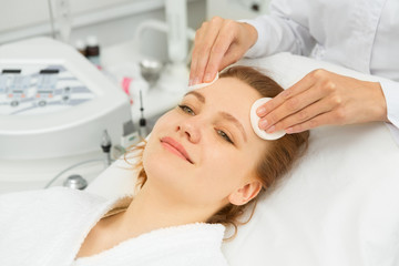 Obraz na płótnie Canvas Happy young beautiful woman smiling joyfully while professional beautician cleansing her face with cotton pads preparing for facials at the beauty spa salon clinical cosmetology dermatology skincare 