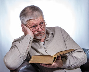 An elderly man with glasses holds a book in his hands but does not read it