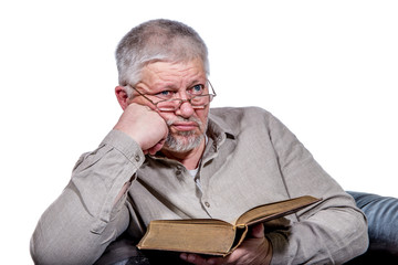 An elderly man with glasses holds a book in his hands but does not read it