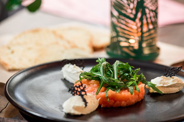 Delicious salmon tartare with toasted bread, cheese and arugula on a black plate in the restaurant. Healthy lunch meal made of raw salmon. Classical French cuisine.